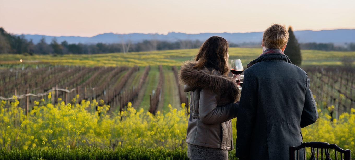 Couple enjoying a romantic sunset dinner overlooking Napa Valley vineyards during winter mustard bloom, with wine glasses and candlelit table on a terrace restaurant.
