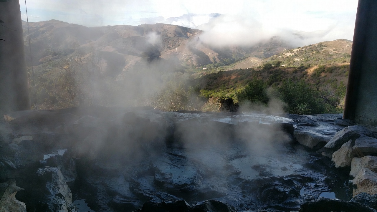Steam rising from a traditional geothermal mud bath in Calistoga, Napa Valley, with natural stone textures and a view of the northern valley foothills. 