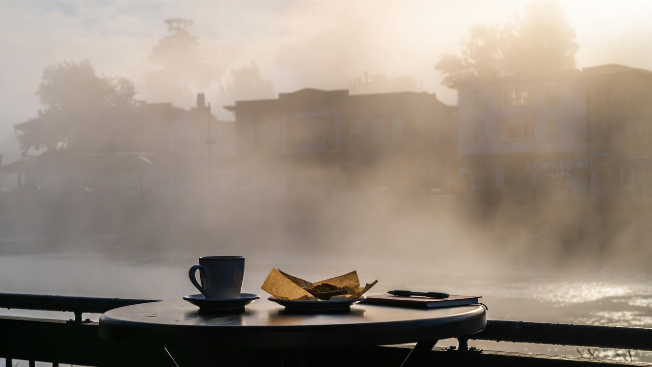 Early morning coffee at an outdoor café in downtown Napa with a ceramic cup and pastry on a table, soft marine fog along the river, capturing a calm Napa Valley coffee route for East Bay visitors.
