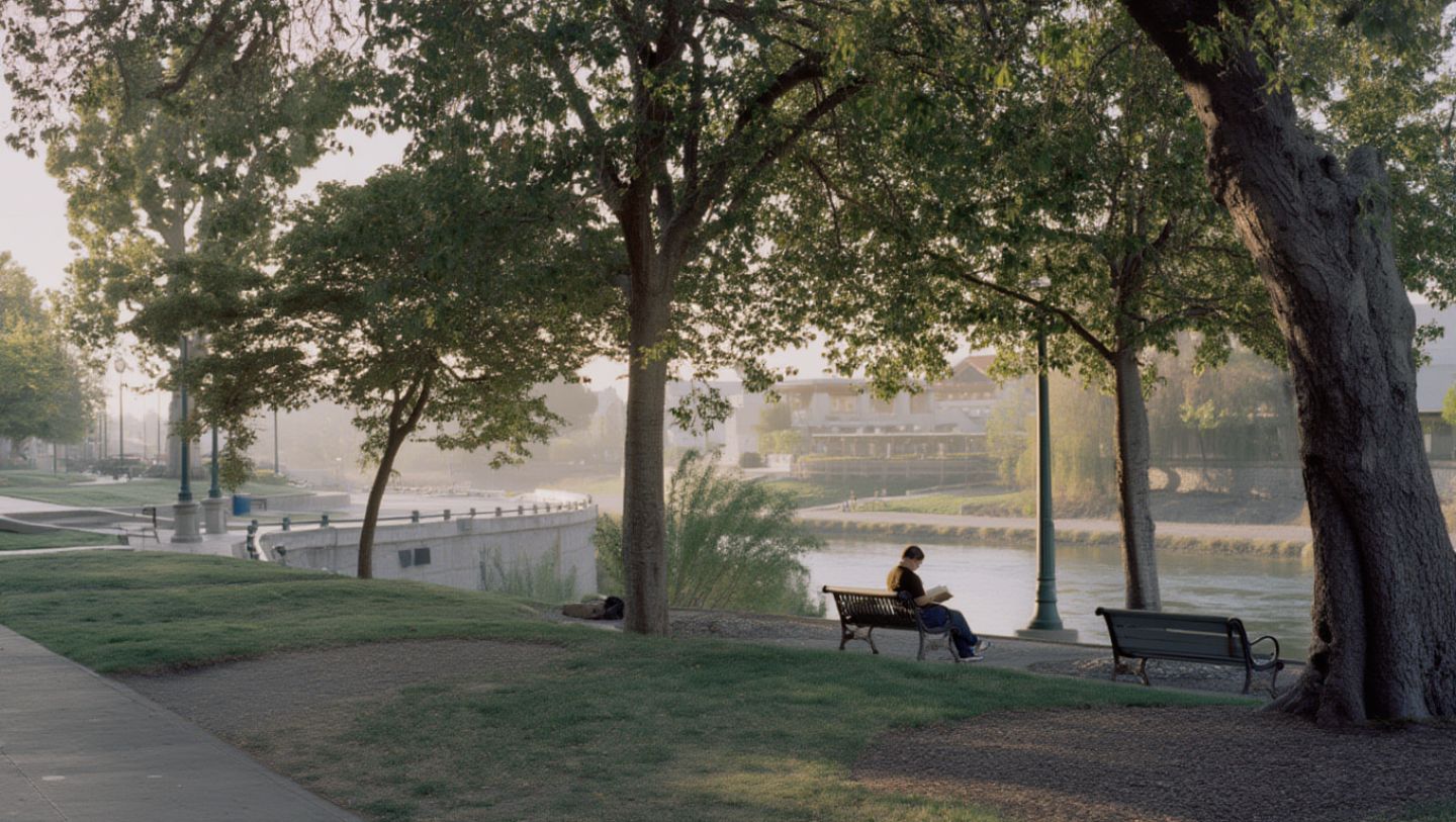 Person reading a book on a shaded bench along the Napa River in downtown Napa with trees and morning light reflecting on the water.