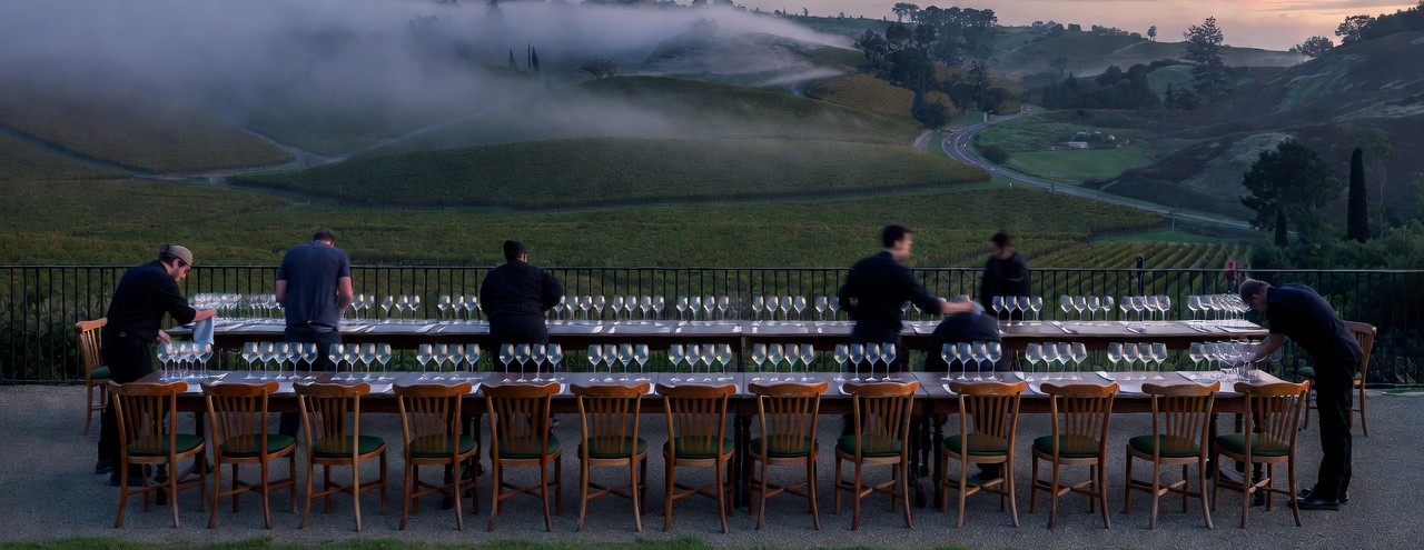 Early morning at a Napa Valley estate along Silverado Trail with staff preparing a tasting terrace, aligning chairs and polishing wine glasses as vineyard rows sit beneath lifting morning fog.