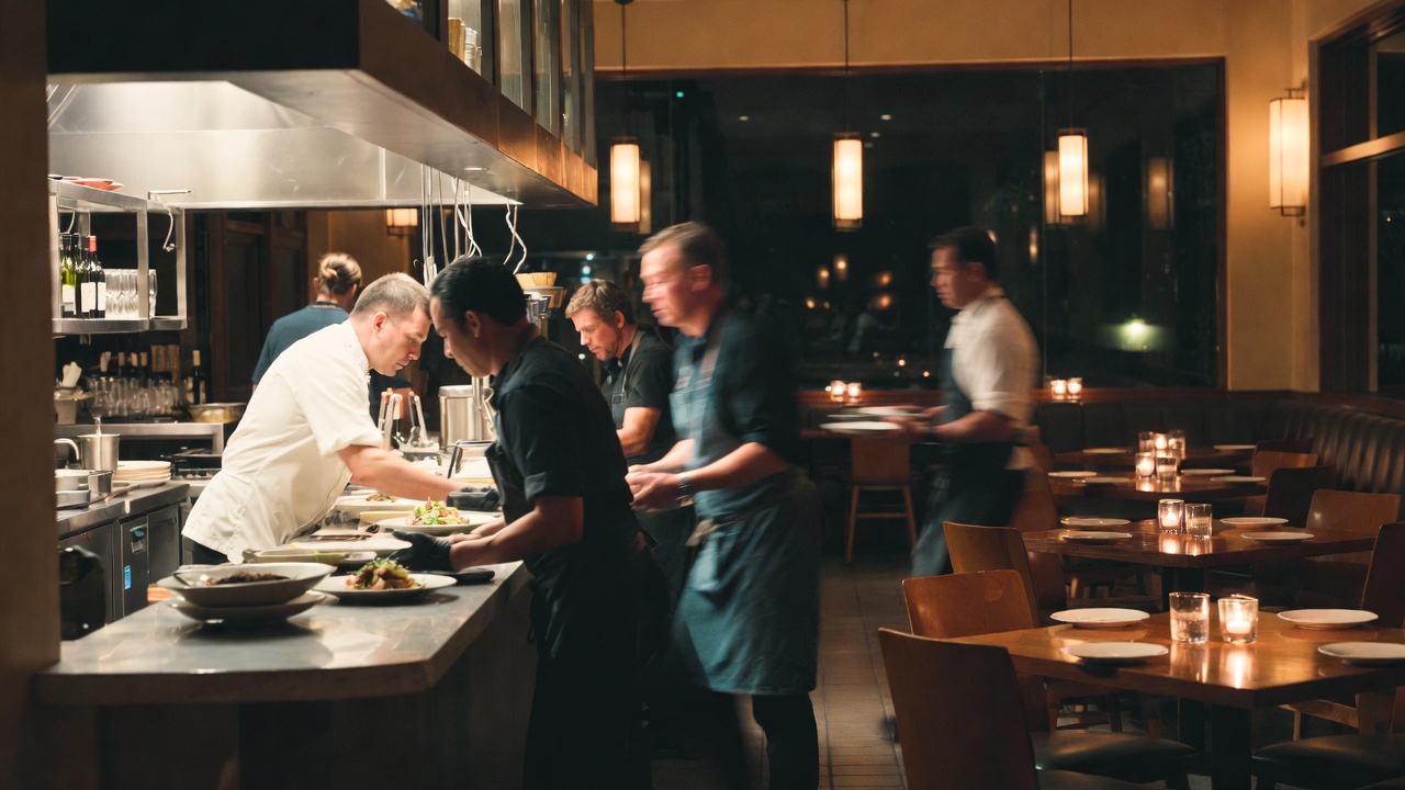 Chef plating seasonal vegetables in a refined Yountville restaurant dining room in Napa Valley while servers prepare for evening service.
